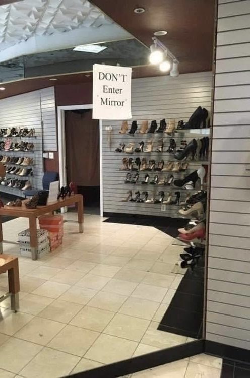A low quality digital photo of a shoe store with a piece of A4 printed paper stuck to the wall mirror that says 'DON'T Enter Mirror'.